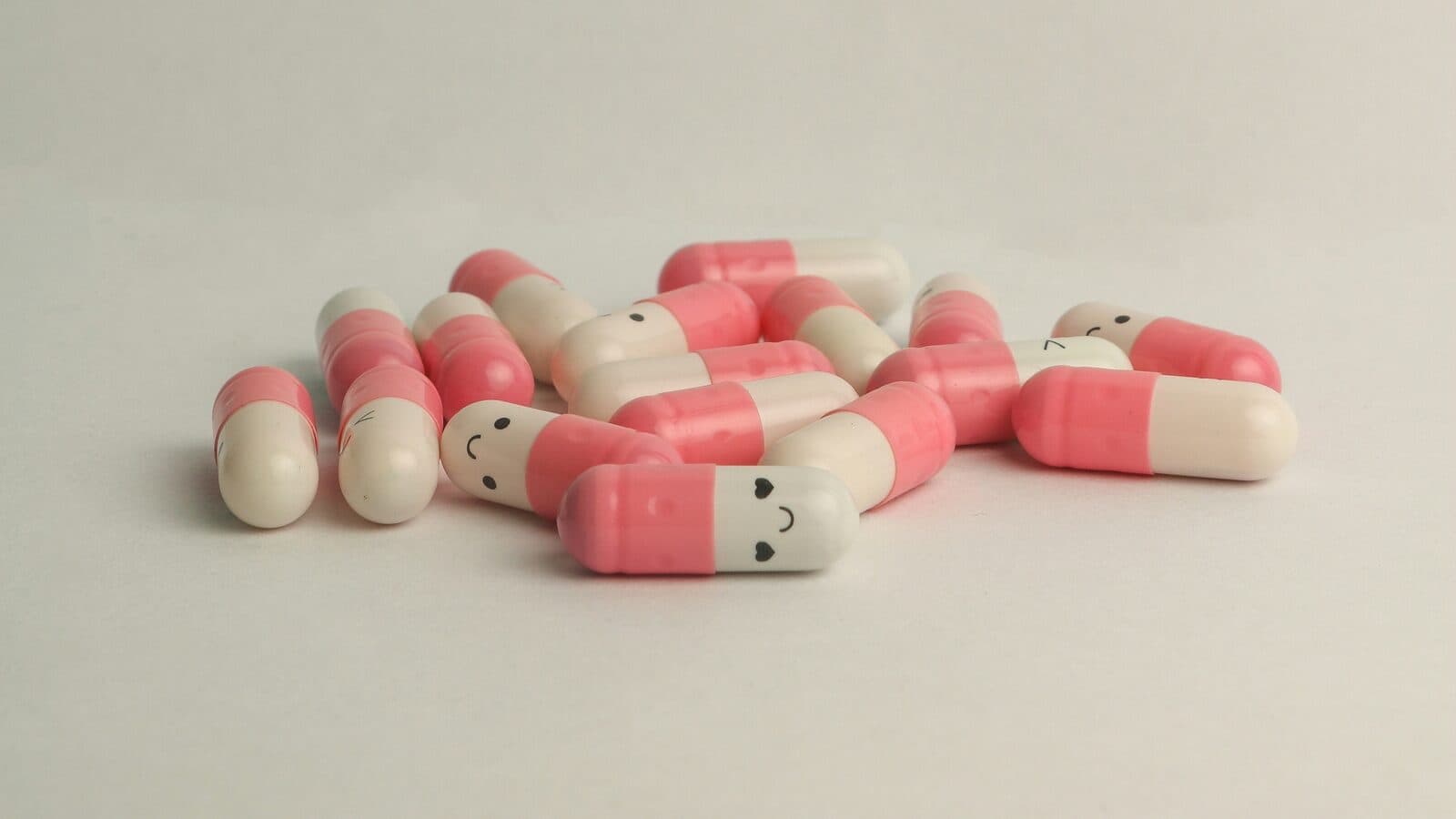 Pink-and-white medicine capsules, some with small drawn faces, scattered on a light background.
